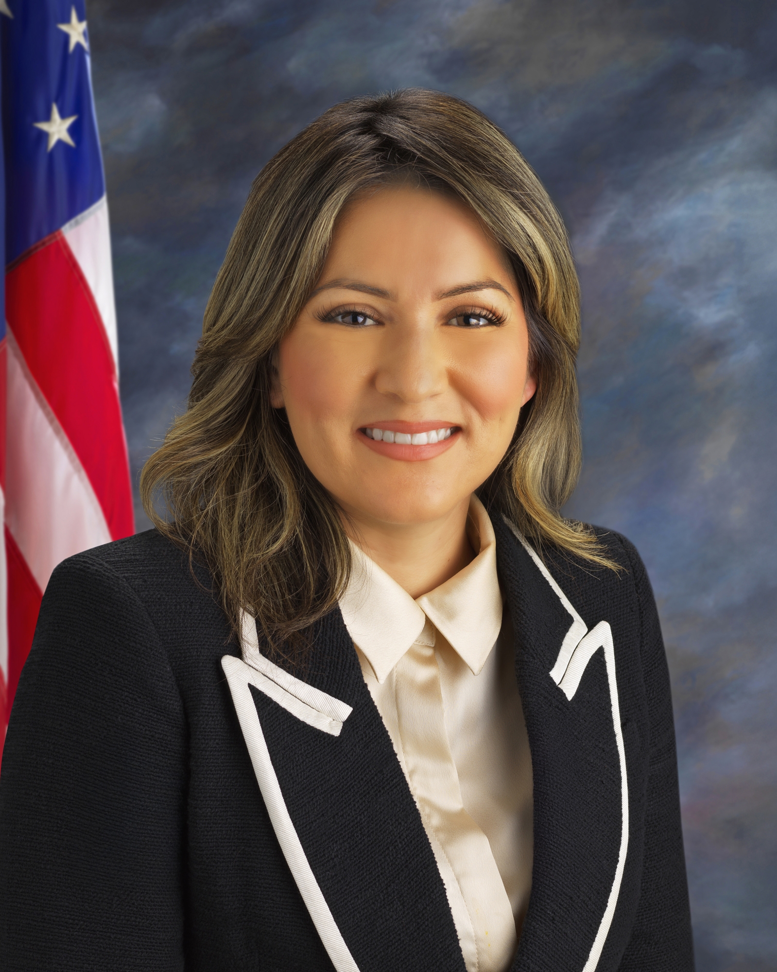 Headshot of Council woman Daisy Macias. Grey background with an American flag on the left side of the photo behind the council member